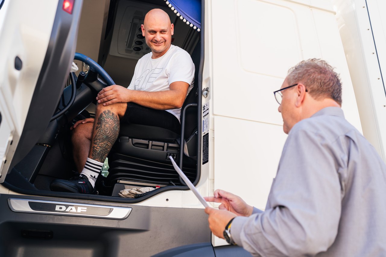 Ein lächelnder LKW-Fahrer im Führerhaus spricht mit einem Mann, der Dokumente hält.