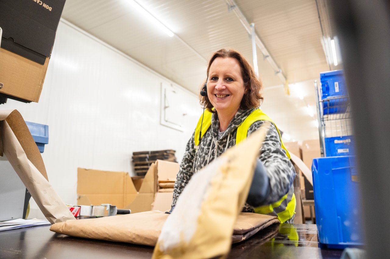 Eine lächelnde Frau in gelber Weste verpackt eine Tüte in braunes Papier.